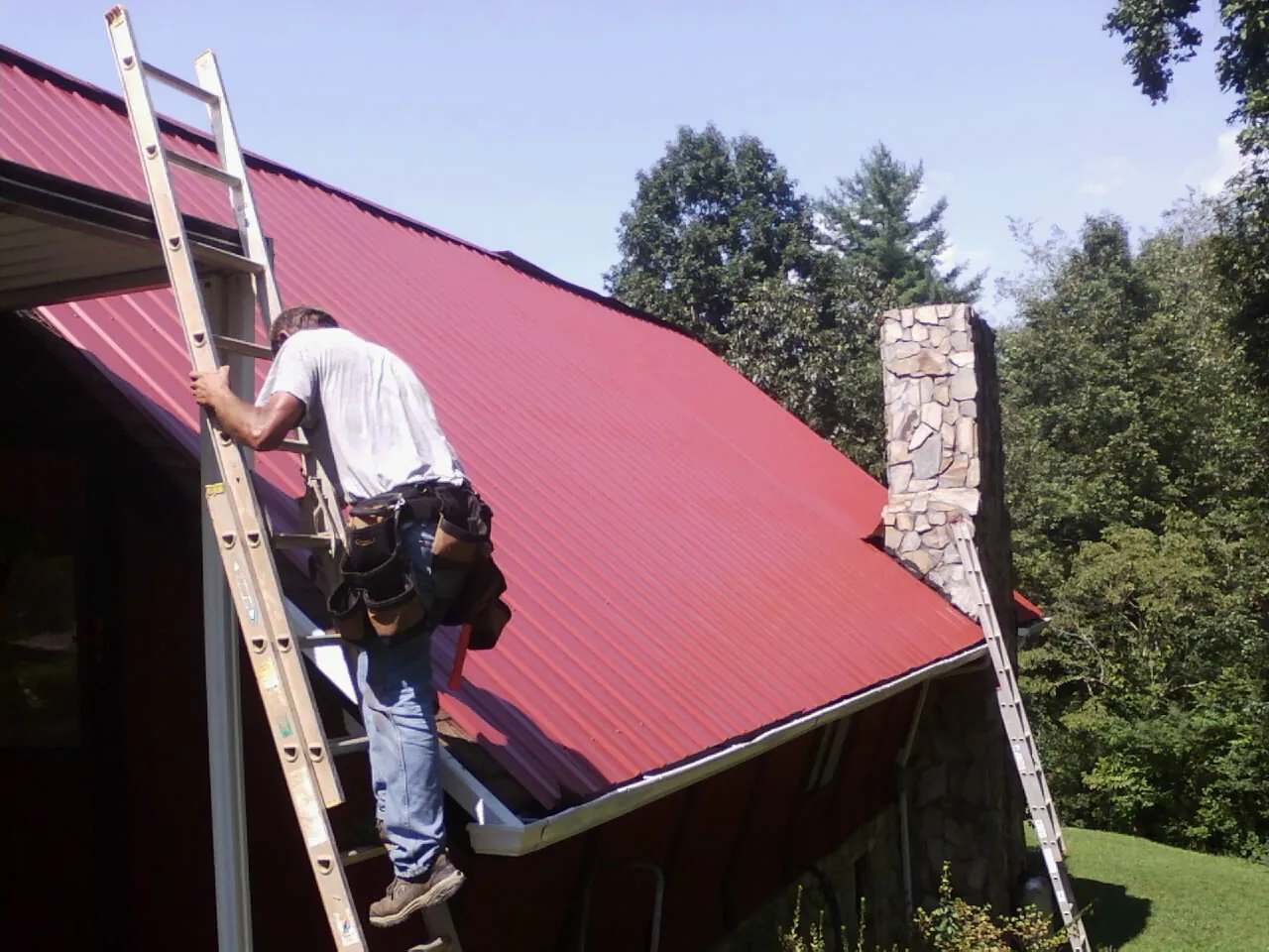 Roofer on a ladder beside a red metal roof and stone chimney — Soffit & Fascia Repair by Roofing Brothers in Nicholson